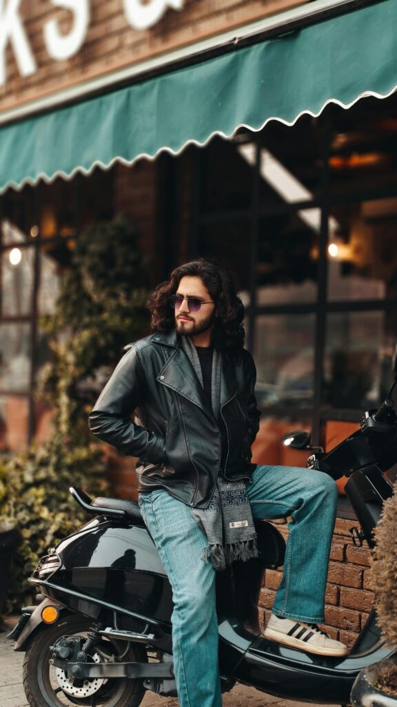 Stylish man with sunglasses and leather jacket sits on a scooter outside a cafe. Urban and casual vibe.
